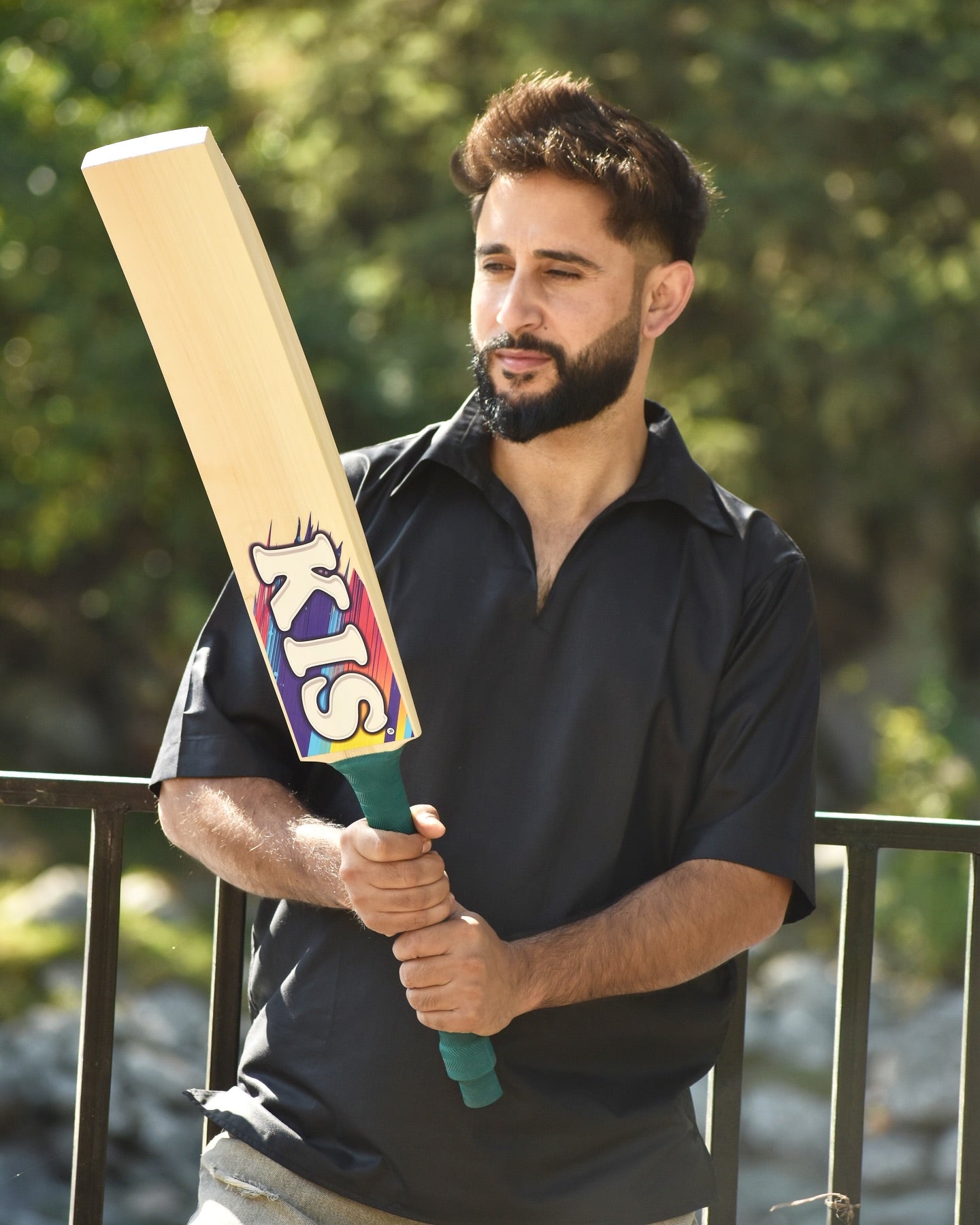 Man holding a cricket bat with 'KIS' branding outdoors